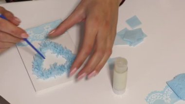 Bir kadın masaya oturur ve tebrik kartı hazırlar. Kağıt elementleri yapıştırır. Yukarıdan çekilen.