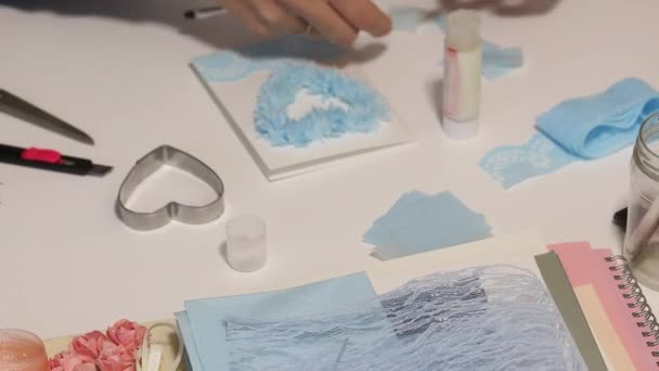 The woman at the table makes a greeting card. Glues paper elements in the shape of a rose. Tools and materials are laid out nearby. Filmed from above — Stock Video