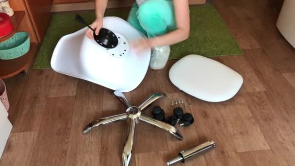 La mujer recoge una silla de ordenador sobre ruedas. Las piezas de la silla de oficina están en el piso del apartamento. — Vídeos de Stock