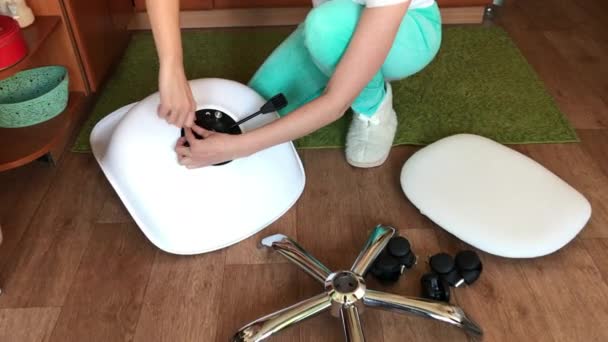 Kadın tekerlekli bir bilgisayar sandalyesi topluyor. Cıvataları sıkılaştırır. Ofis sandalyesinin parçaları dairenin zemininde.. — Stok video