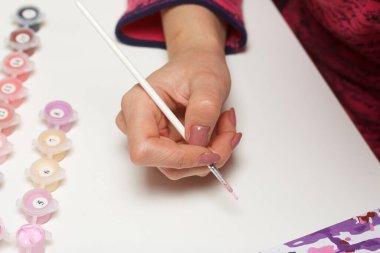 Kadın sayılarla resim yapıyor. Tuval üzerinde akrilik boya. Yakınlarda boya kutuları ve diğer aksesuarlar var..