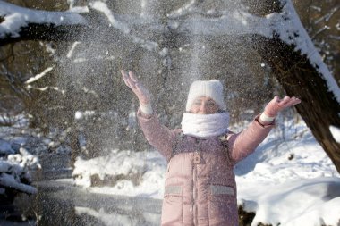 Parkta yürüyüş yapan kışlık elbiseli bir kadın. Bir avuç dolusu kar kusuyor. Yakınlarda bir nehir akıyor. Etrafta çok kar var..