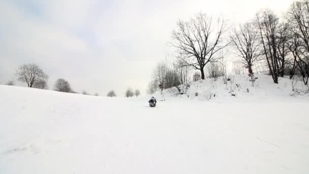 La niña sube a una colina cubierta de nieve, se sienta en un trineo y se desliza hacia abajo hasta el fondo de la colina. Voltea la cabeza sobre los talones, el tablero se desliza. Rodando la cabeza sobre los talones, acostado en la nieve. — Vídeos de Stock