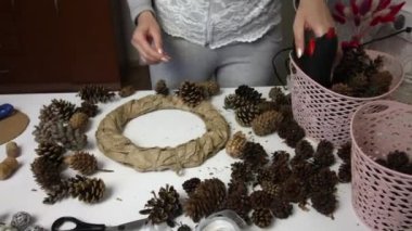 Bir kadın çam kozalağından çelenk yapar. Karton ve kağıttan yapılmış bir yüzüğe koni takıyor. Koniler yakınlardaki masa yüzeyine yayılmış durumda..