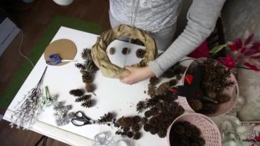 Bir kadın çam kozalağından çelenk yapar. Karton bir yüzüğe kağıt takıyor. Yukarıdan çekilmiş. Koniler yakınlardaki masa yüzeyine yayılmış durumda..