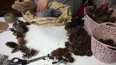 Bir kadın çam kozalağından çelenk yapar. Karton bir yüzüğe kağıt takıyor. Tutkal tabancası kullanıyor. Koniler yakınlardaki masa yüzeyine yayılmış durumda.