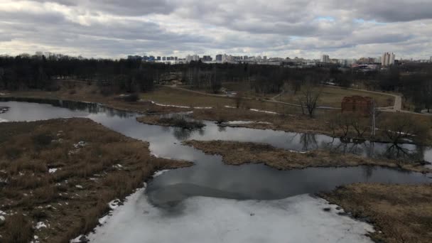 Flight over the river in the spring park. Remnants of snow are visible in places. Spring landscape. Multi-storey buildings are visible on the horizon. — Stock Video