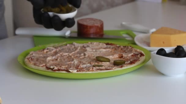 Um homem coloca fatias de pimenta de espaguete em uma base de pizza espalhada com pasta de tomate. Os ingredientes de cozinha são colocados nas proximidades. A fazer pizza em casa. Imagem de close-up. — Vídeo de Stock