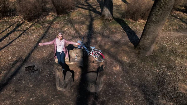 Girl on a bike ride walk in the spring park. Sits on a bench and looks at the phone. Her bike is nearby. Aerial photography taken with a drone.