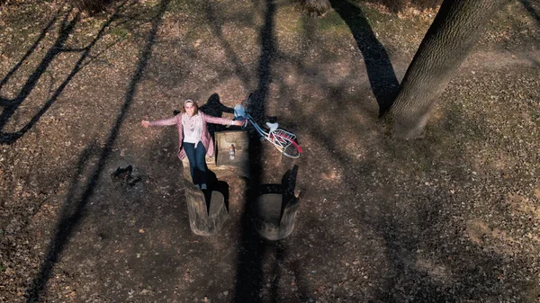 Girl on a bike ride walk in the spring park. Sits on a bench and looks at the phone. Her bike is nearby. Aerial photography taken with a drone.