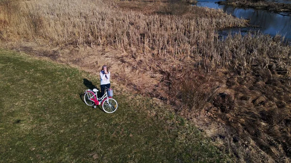 Girl on a bike ride walk in the spring park. Stands on the lawn in the park. She is holding a bicycle. Aerial photography taken with a drone.