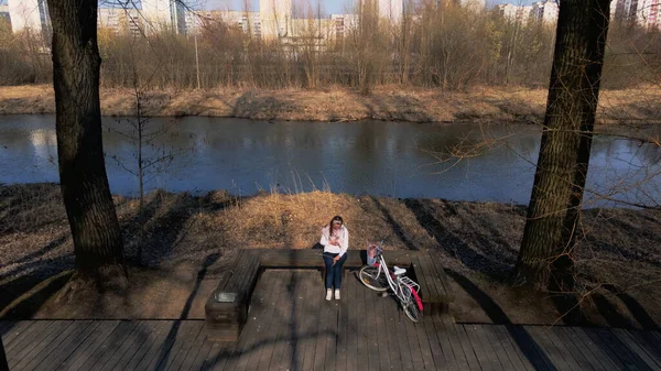 Girl on a bike ride walk in the spring park. Sits on a bench and looks at the phone. Her bike is nearby. Aerial photography taken with a drone.