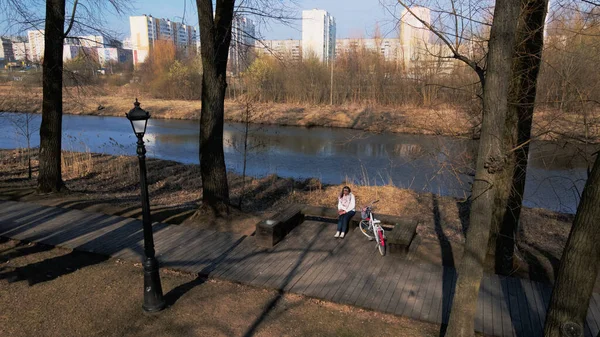 Girl on a bike ride walk in the spring park. Sits on a bench and looks at the phone. Her bike is nearby. Aerial photography taken with a drone.