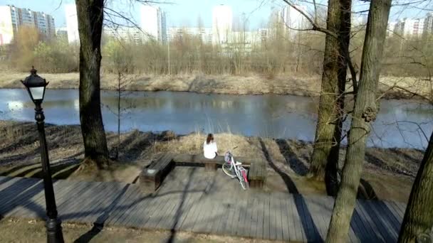 Dívka sedí na lavičce u řeky v jarním parku. Nedaleko je kolo.. — Stock video