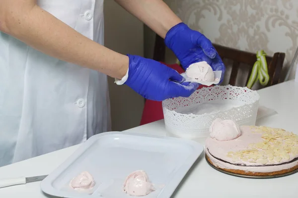 A woman prepares a marshmallow cake. Makes roses from marshmallows for decoration and places on the cake. Works with a mask and gloves.