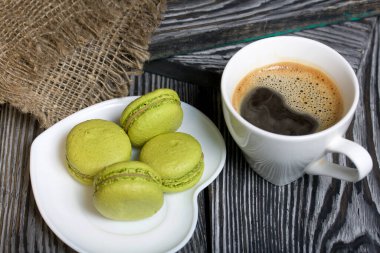 Açık yeşil badem kurabiyeleri bir çay tabağının üzerinde. Yanında bir fincan kahve ve keten kumaş var..