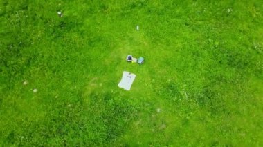 Yeşil bir çayırda güneşlenen bir kadın. Sarı bir halıda, karnının üstünde yatıyordu. İnsansız hava aracı onun üzerinde daireler çiziyor. Hava fotoğrafçılığı.