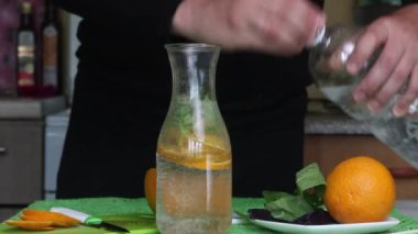 Bir adam şişeye portakal ve fesleğen koyar. Suyu döker ve kapağı kapatır. Turuncu ve fesleğenli su.