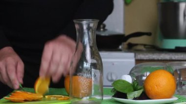 Adam portakal kesiyor. Portakal ve fesleğeni şişeye koyar. Masanın yanında bir şişe su var. Turuncu ve fesleğenli su.