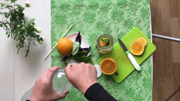 Un hombre pone una naranja y albahaca en una botella. Vierte agua. Agua infundida con naranja y albahaca. Recetas de agua de desintoxicación. Filmado desde arriba. — Vídeos de Stock