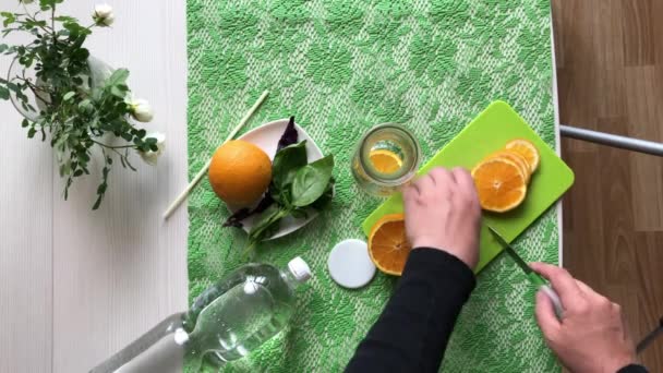 Un uomo mette un'arancia e del basilico in una bottiglia. Acqua infusa con arancia e basilico. Ricette acqua Detox. Girato dall'alto. — Video Stock