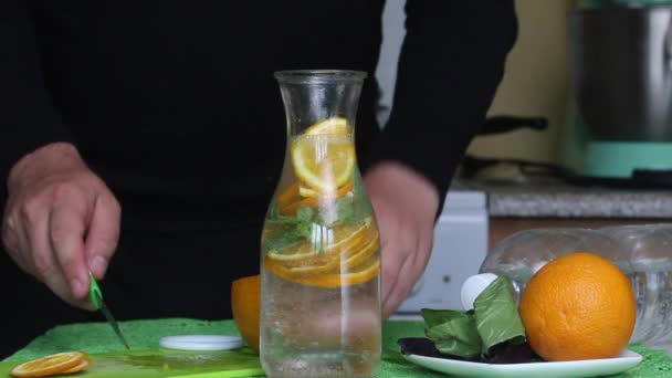 Un homme coupe une orange et la met dans une bouteille d'eau. Eau infusée d'orange et de basilic. Recettes d'eau detox. — Video