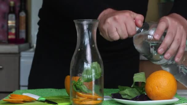 Un homme met une orange et du basilic dans une bouteille. Verse de l'eau. Eau infusée d'orange et de basilic. — Video