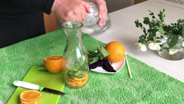 一个男人把一个橙子和罗勒放进瓶子里.倒水。注入橙子和罗勒的水。排毒水配方。从侧面开枪 — 图库视频影像