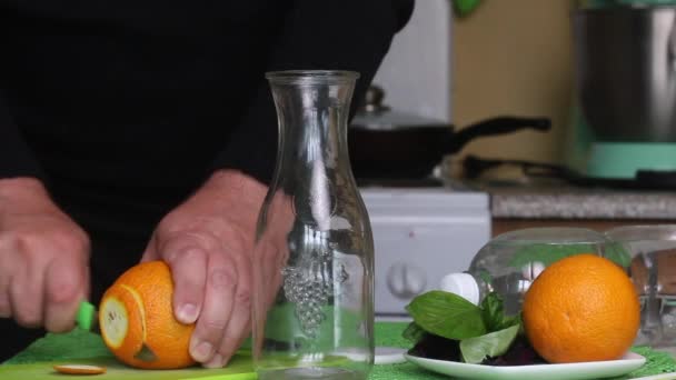 O homem está a cortar uma laranja. Manjericão e água estão sobre a mesa. Água infundida com laranja e manjericão. Desintoxicação receitas de água. — Vídeo de Stock