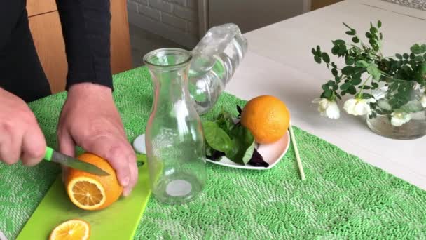 Ο άνθρωπος κόβει ένα πορτοκάλι. Ο Μπάζιλ και το νερό είναι στο τραπέζι. Νερό εμποτισμένο με πορτοκάλι και βασιλικό. Συνταγές αποτοξίνωσης νερού. Πυροβολήθηκε από το πλάι. — Αρχείο Βίντεο