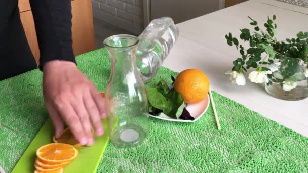 Un homme met une orange et du basilic dans une bouteille. Verse de l'eau. Eau infusée d'orange et de basilic. Recettes d'eau detox. — Video