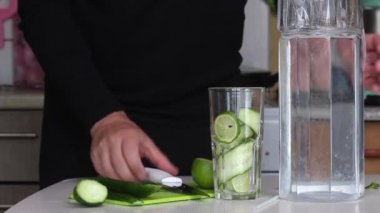 Bir adam dilimlenmiş kireç ve salatalığı bardağa koyar. Onları bir sürahide suyla dolduruyor. Limon ve salatalık ile karıştırılmış pişirme suyu. Detoks su tarifi. Yakın çekim..