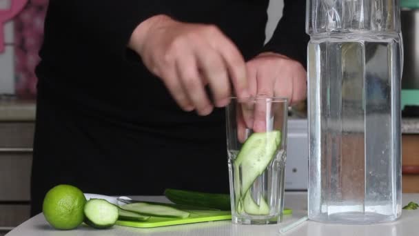 Um homem corta um pepino e coloca-o num copo. Limão próximo e água em um decantador. Água de cozinha infundida com limão e pepino. Receita de água desintoxicada. Imagem de close-up. — Vídeo de Stock