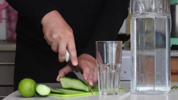 Un homme coupe un concombre. A proximité de chaux et d'eau dans une carafe. Eau de cuisson infusée de chaux et de concombre. Recette d'eau de désintoxication. Gros plan — Video