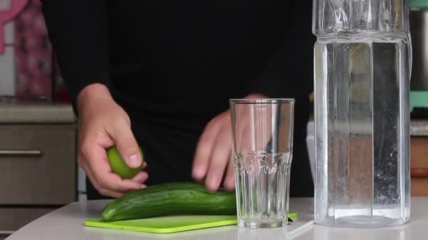 Een man snijdt een komkommer. Vlakbij limoen en water in een karaf. Kookwater doordrenkt met kalk en komkommer. Detox water recept. Close-up opname. — Stockvideo