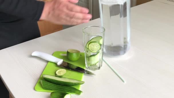 Un homme met de la chaux tranchée et du concombre dans un verre. Remplissez-les avec de l'eau d'une carafe. Eau de cuisson infusée de chaux et de concombre. Recette d'eau detox. — Video