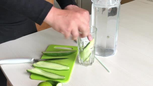 Een man snijdt een komkommer en doet hem in een glas. Vlakbij limoen en water in een karaf. Kookwater doordrenkt met kalk en komkommer. Detox water recept. — Stockvideo