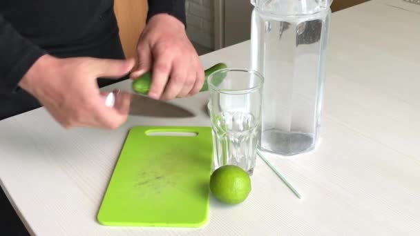 Een man snijdt een komkommer. Vlakbij limoen en water in een karaf. Kookwater doordrenkt met kalk en komkommer. Detox water recept. — Stockvideo