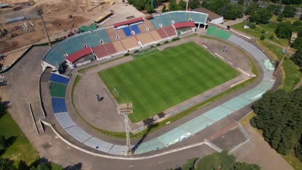 Foci stadion a városi parkban. Egy új aréna épül a közelben. A zöld mező és állványok láthatók, festett különböző színekben. Oldalról repül. Repülj visszafelé. Légi fényképezés. — Stock videók