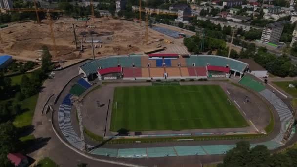 Football stadium in the city park. A new arena is being built nearby. A green field and stands are visible, painted in different colors. Aerial photography. — Video
