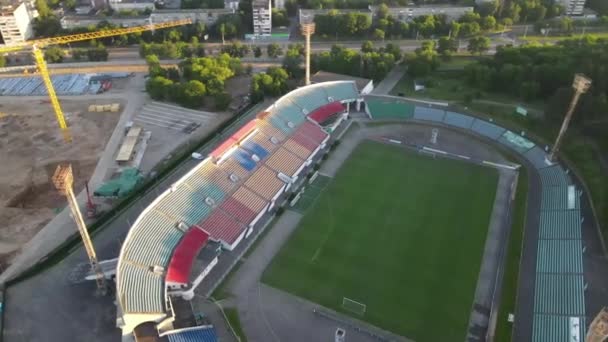 市内公園内のサッカースタジアム。緑のフィールドとスタンドが表示され、異なる色で塗装されます。上昇する太陽の光の中で弧を描いて飛んでいます。空中写真. — ストック動画
