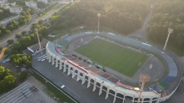 市内公園内のサッカースタジアム。緑のフィールドとスタンドが表示され、異なる色で塗装されます。上昇する太陽の光線の中で円を描いて飛んでいます。空中写真. — ストック動画