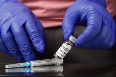 Koronavirüse karşı aşı. Aşısı olan bir ampulün ellerinde. Masanın üzerinde enjeksiyon için bir şırınga var. Eldivenli eller. Yakın çekim..