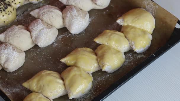 Una donna spalma un uovo su un bianco per il pane, spruzza con semi di sesamo. Pane a forma di spillo si trova su una teglia da forno. Primo piano.. — Video Stock