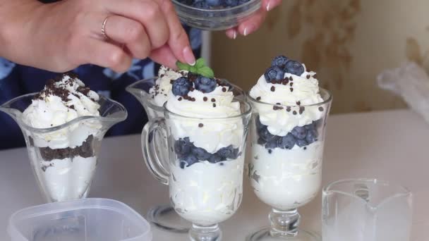 Uma mulher prepara uma sobremesa com chantilly, mirtilos e chocolate. Decora a sobremesa com folhas de hortelã. Imagem de close-up. — Vídeo de Stock