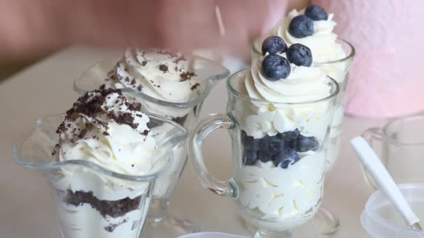 Uma mulher prepara uma sobremesa com chantilly, mirtilos e chocolate. Decora a sobremesa com mirtilos e chocolate. Ao lado dos ingredientes para a sobremesa. — Vídeo de Stock