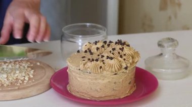 A woman chops peanuts and sprinkles on a carrot cake decorated with cream and chocolate drops. Close-up shot.