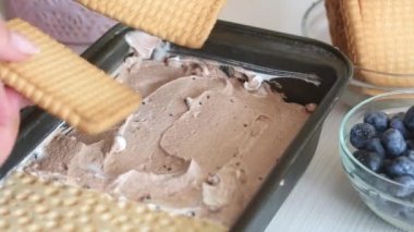 The woman places the cookies on top of the cream in the container. Makes ice cream from cream, biscuits and crispbread. Other ingredients are spread out on the table nearby. Close-up shot.