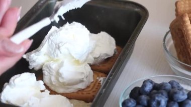A woman adds whipped cream to a container on top of a cookie. Makes ice cream from cream, biscuits and crispbread. Other ingredients are spread out on the table nearby. Close-up shot.