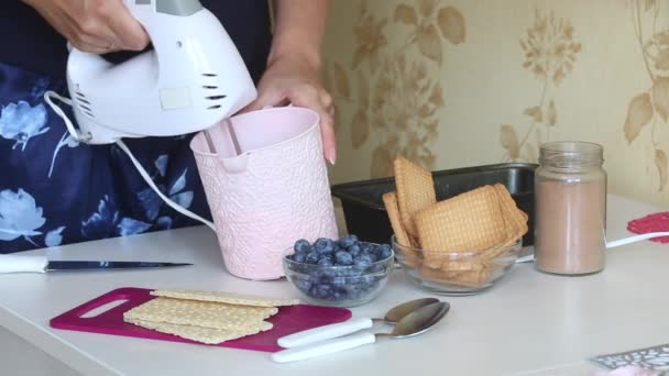 Uma mulher está a fazer chantilly num recipiente. Faz sorvete de creme, biscoitos e pão crocante. Os ingredientes estão localizados ao lado uns dos outros na mesa. — Vídeo de Stock